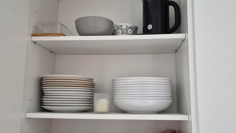 An open kitchen cabinet with various dishware. On the top are cups and a coffee machine, while below, plates and bowls are neatly organized.
