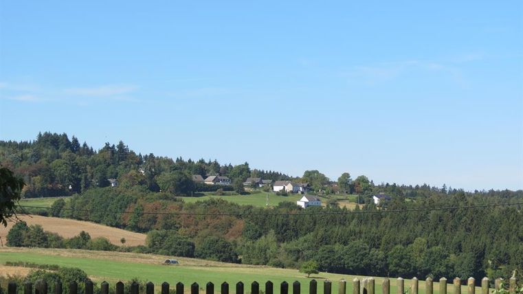 Een rustig landschap met glooiende heuvels en hier en daar een huis. De lucht is helder en blauw, omringd door groene bossen en velden.