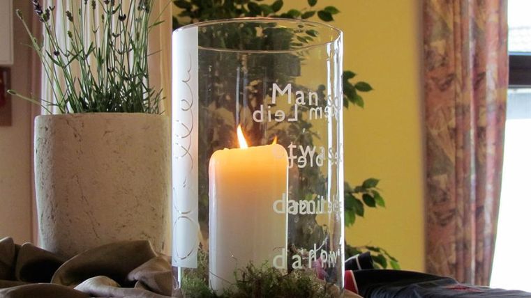 A burning candle in a glass container stands on a table. In the background, plants and decorative fabrics can be seen.