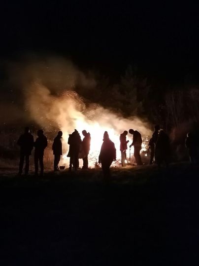 Eine Gruppe von Menschen steht in der Nacht um ein großes Feuer. Der Rauch steigt auf und die Silhouetten der Personen sind sichtbar.