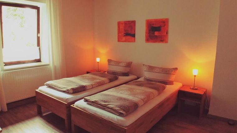 A cozy bedroom with two single beds and warm light. Two colorful pictures hang on the wall, and a window lets in daylight.