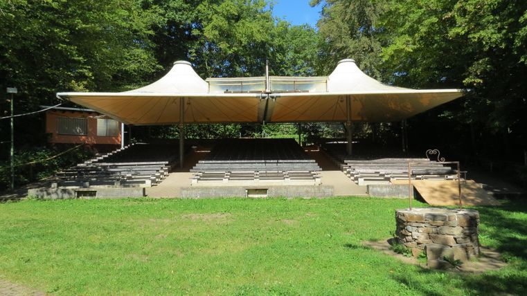 Eine Freiluftarena mit einem markanten, überspannten Dach und hölzernen Sitzplätzen. Die Umgebung ist von Bäumen umgeben und der Himmel ist klar und blau.