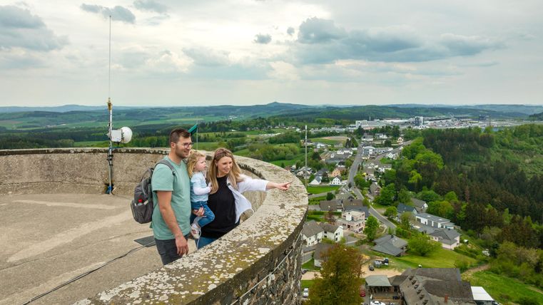 Eine Familie steht auf einem Aussichtsturm und blickt auf die Landschaft. Im Hintergrund sind grüne Hügel und kleine Häuser zu sehen.