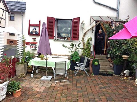 Een uitnodigende binnenplaats met een tafel en stoelen onder een paarse parasol. Op de achtergrond staat een persoon voor de voordeur en enkele planten zijn verspreid over de ruimte.