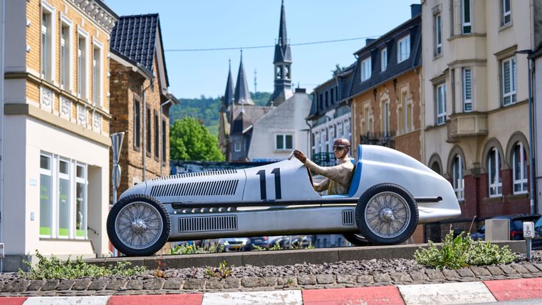 Statue eines Rennwagens mit Fahrer in einer Stadt mit historischen Gebäuden im Hintergrund.