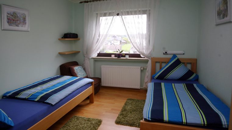 Een gezellige kamer met twee eenpersoonsbedden en kleurrijke dekbedden. Er is een stoel, een tapijt en grote ramen met gordijnen.