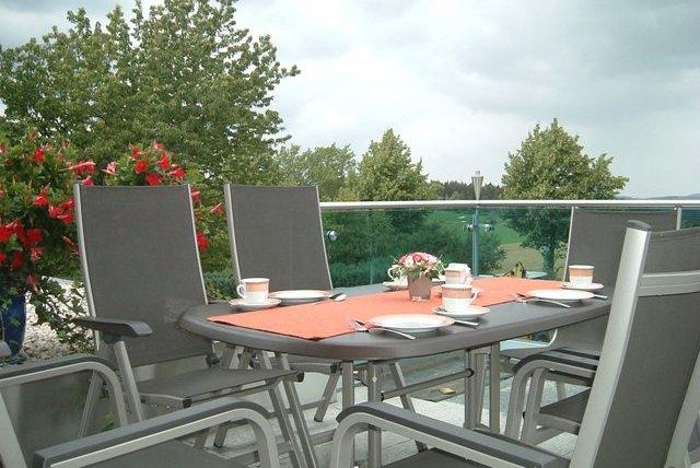 Ein gemütlicher Tisch auf einer Terrasse mit Stühlen und einer Tischdecke. Im Hintergrund sind grüne Bäume und blühende Blumen zu sehen.