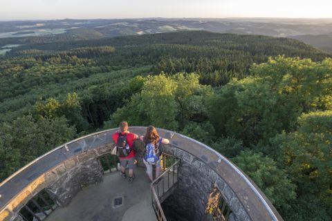 Ein Aussichtsturm inmitten einer grünen Landschaft. Zwei Wanderer genießen die Aussicht auf die umliegenden Wälder und Felder.