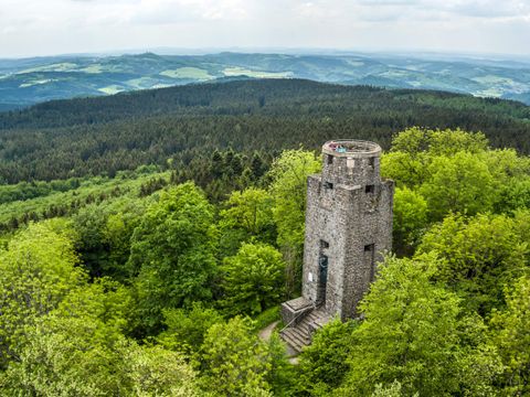 Ein steinerner Aussichtsturm umgeben von dichtem Grün. Im Hintergrund erstrecken sich sanfte Hügel und Wälder.