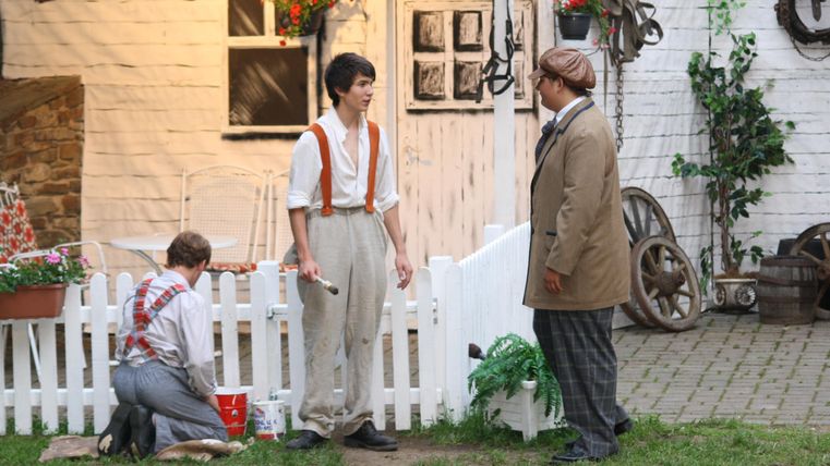 Drei Männer stehen vor einem weißen Zaun und einem Bauernhaus. Einer von ihnen färbt einen Zaun, während die anderen miteinander sprechen.