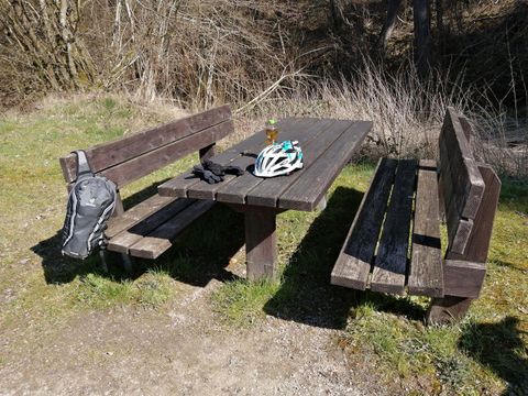 Ein Holzplatz mit einer Bank und einem Tisch im Freien. Auf dem Tisch liegen ein Fahrradhelm, Kleidung und eine Flasche.