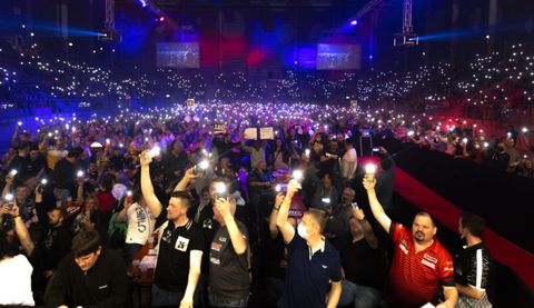 Eine große Menschenmenge bei einem Event, die ihre Handys mit eingeschaltetem Licht in die Höhe hält. Bunte Lichter und eine festliche Atmosphäre prägen die Szene.