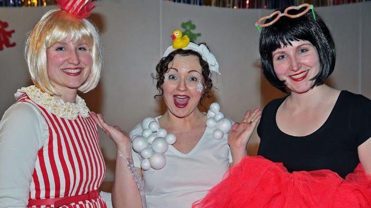Drie vrouwen in kleurrijke kostuums staan glimlachend bij elkaar. De eerste draagt een popcornkostuum, de tweede is verkleed als zeepbel en de derde draagt een zwarte jurk met rode tule.