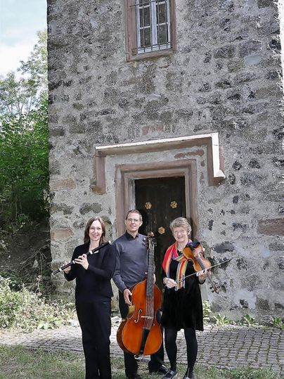 Eine Gruppe von drei Musikern steht vor einem alten Gebäude. Sie spielen Flöte, Cello und Geige in einer grünen Umgebung.