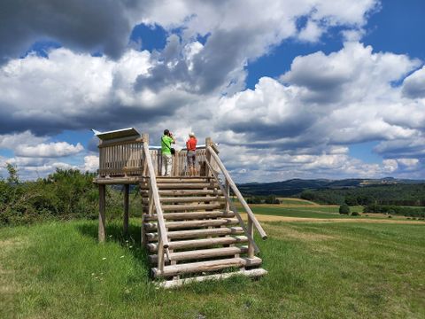 Zwei Personen stehen auf einer hölzernen Aussichtsplattform in einer ländlichen Landschaft mit bewölktem Himmel.
