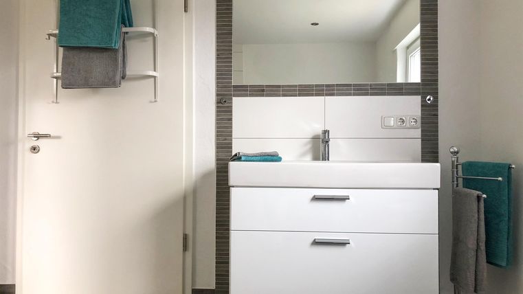 Een moderne badkamer met een witte wastafel en een grote spiegel. Handdoeken in blauw en grijs zijn aan de muur opgehangen.