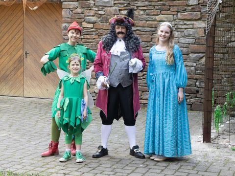 Eine Gruppe von Personen in bunten Kostümen steht vor einer Steinmauer. Sie stellen Charaktere aus einem Märchen dar, darunter ein Junge in einem grünen Kostüm und ein Kapitän in einem roten Cape.