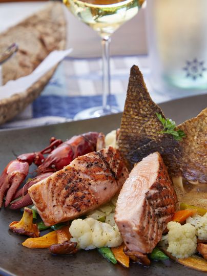 Ein köstlicher Fischgericht mit gegrilltem Lachs, buntem Gemüse und knackigen Garnelen. Im Hintergrund ein Gläschen Weißwein.