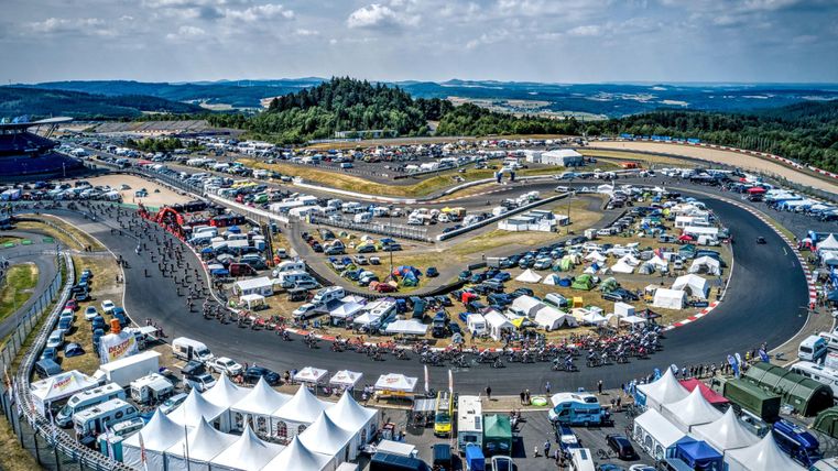 Eine breite Rennstrecke mit vielen Kurven und Zuschauerbereichen. In der Umgebung sind zahlreiche Autos und Zelte zu sehen.