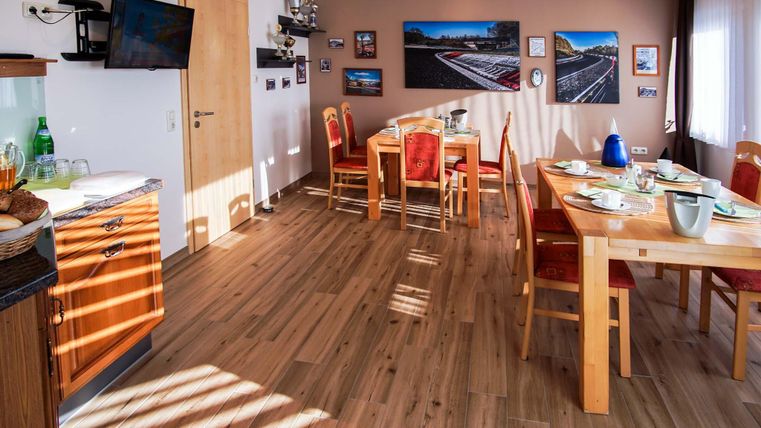 Ein helle und einladende Wohnküche mit einem großen Tisch und roten Stühlen. An den Wänden hängen Bilder, und der Boden ist aus hellem Holz.