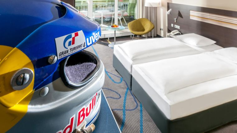 A modern hotel room with two beds and a cozy seating area. In the foreground is a Red Bull racing helmet.