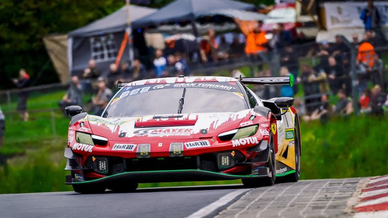 Ein Rennwagen fährt auf einer Rennstrecke. Im Hintergrund sind Zuschauer und Zeltstrukturen zu sehen.