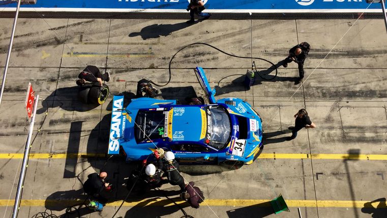 Ein Rennwagen in der Boxengasse wird von einem Team gewartet. Mechaniker arbeiten an Reifen und Technik.