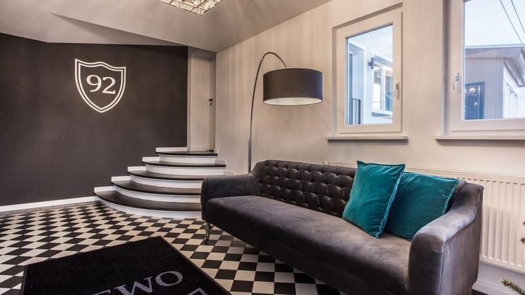 Ein modernes Foyer mit einem hellen, eleganten Design. Ein schwarzes Sofa und dekorative Kissen stehen auf einem schwarz-weißen Schachbrettboden.