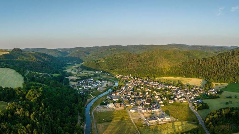 A picturesque view of a small village, surrounded by green hills and forests. A river meanders through the landscape with fields in the background.
