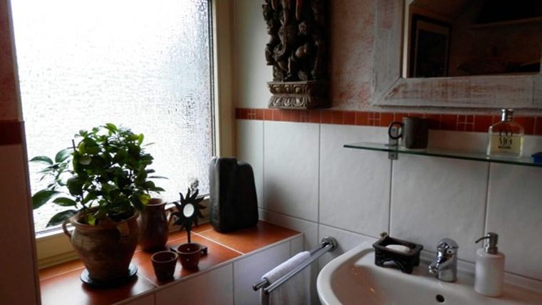 Een gezellige badkamer met een wastafel en een plant op de vensterbank. Het licht valt door een versierd raam en creëert een welkom gevoel.