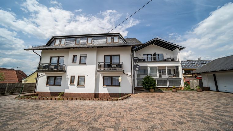 Een modern huis met meerdere balkons en een geplaveide oprit. De lucht is bewolkt, maar het is licht en vriendelijk.