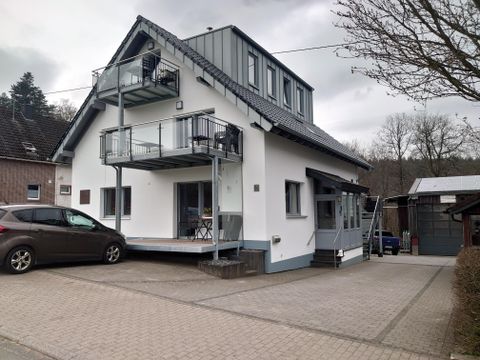 Une maison moderne avec deux balcons et un parking pavé devant la porte. À l'arrière-plan, on peut voir des arbres et un quartier paisible.