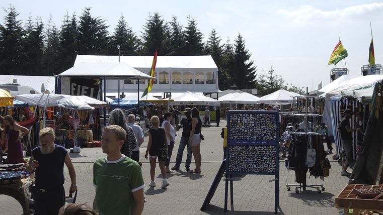 A bustling market with many people and stalls. The tents offer various goods, and some flags can be seen in the background.