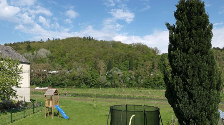 Eine grüne Wiese mit einem Trampolin und einer Rutsche. Im Hintergrund sind Bäume und sanfte Hügel unter einem blauen Himmel zu sehen.