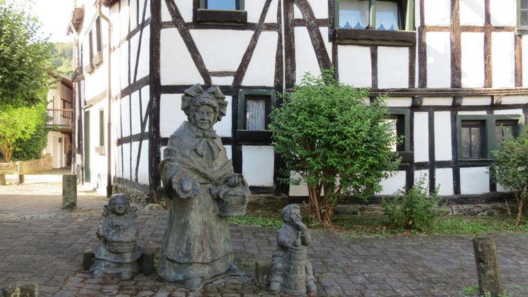 Eine Statue von einer älteren Frau steht auf einem gepflasterten Platz, umgeben von zwei kleineren Figuren. Im Hintergrund ist ein traditionelles Fachwerkhaus zu sehen.