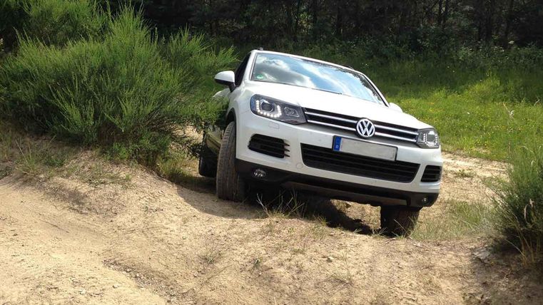 Ein weißes Auto fährt über einen unebenem Weg in der Natur. Im Hintergrund sind grüne Pflanzen und ein klarer Himmel zu sehen.