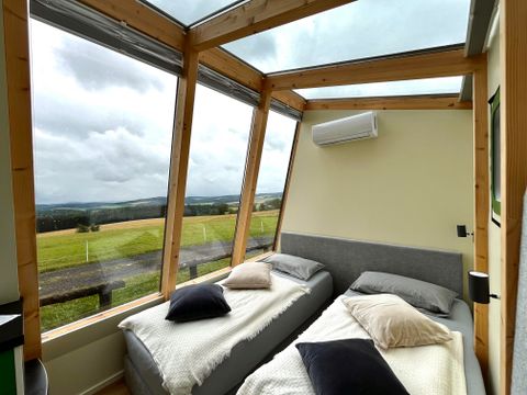 Une chambre confortable avec deux lits et de grandes fenêtres. La vue s'étend sur un paysage verdoyant sous un ciel nuageux.