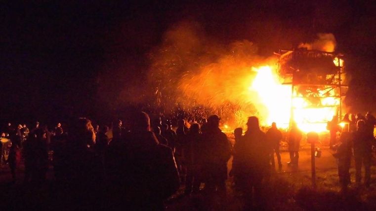 Een grote menigte kijkt naar een vlammend vuur in de nacht. Vonken vliegen door de lucht van een brandend gebouw.