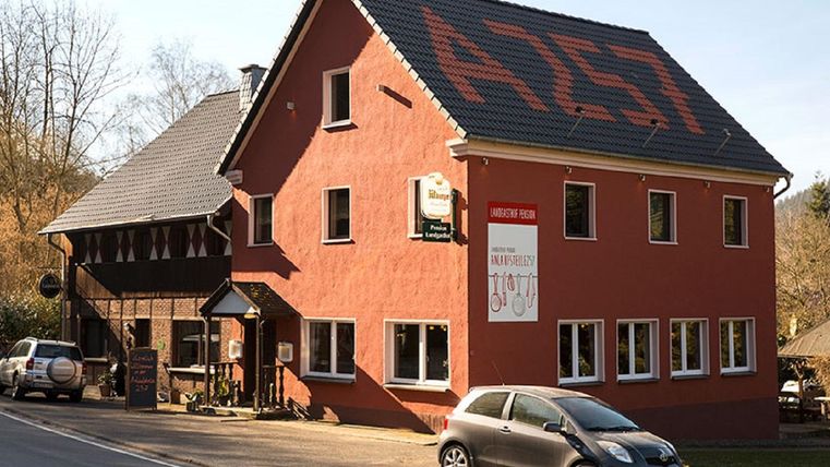 A red building with several windows and a dark roof. In front of it stands a gray car and there is a sign with an inscription.
