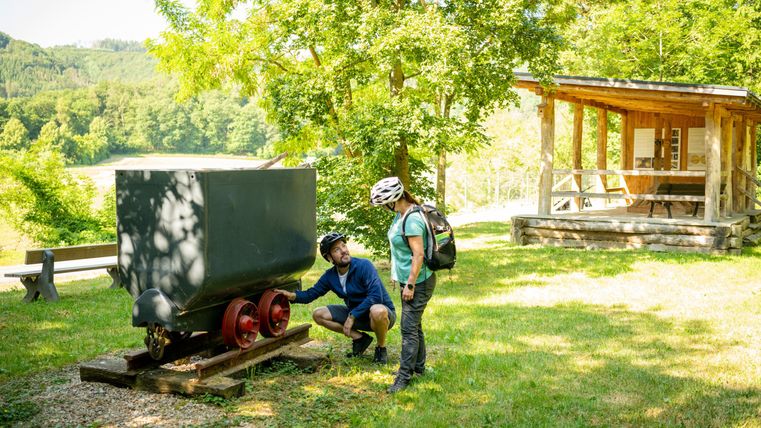 Zwei Radfahrer stehen neben einem historischen Wagen in einer grünen Umgebung. Im Hintergrund ist ein Holzgebäude zu sehen, umgeben von Bäumen.