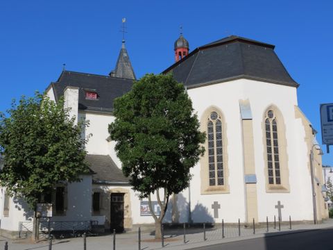 Eine historische Kirche mit hohen Fenstern und einem modernen Anbau. Im Vordergrund steht ein großer Baum.