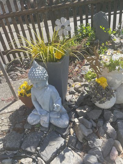 A Buddha statue sits on stones among blooming plants and pots. Surrounded by a well-kept garden, the atmosphere is calm and inviting.