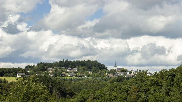 Un paysage calme avec un petit village, entouré de forêts et de collines. Le ciel est couvert de grands nuages blancs.