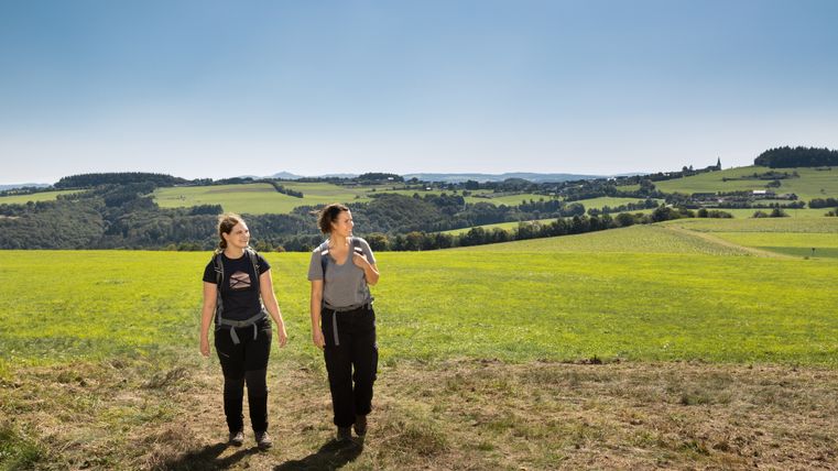 Zwei Frauen wandern auf einem Feldweg mit grünen Hügeln im Hintergrund.