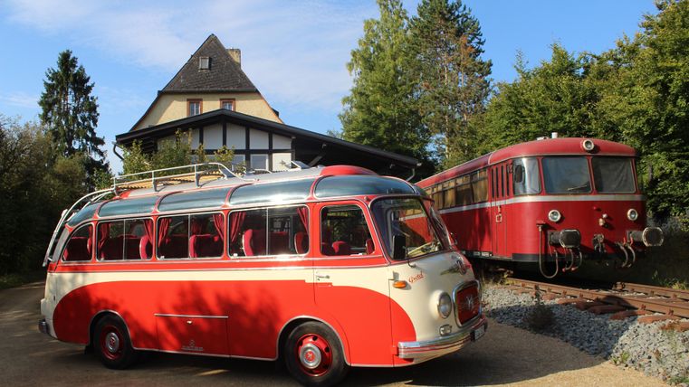 An old red and cream-colored coach stands next to a red locomotive on a track. In the background, there is a house with a pointed roof visible.