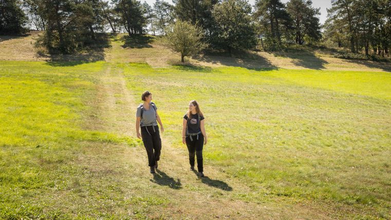 Zwei Frauen gehen auf einem grünen Feld. Im Hintergrund sind Bäume und sanfte Hügel zu sehen.