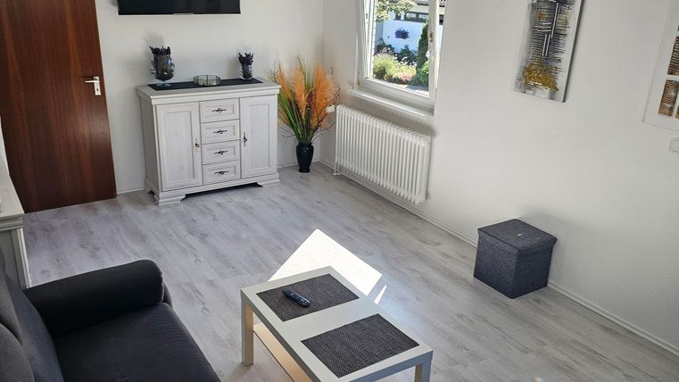 A modern living room with a black sofa and a white table. The room has a bright atmosphere with a television on the wall and decorative plants.