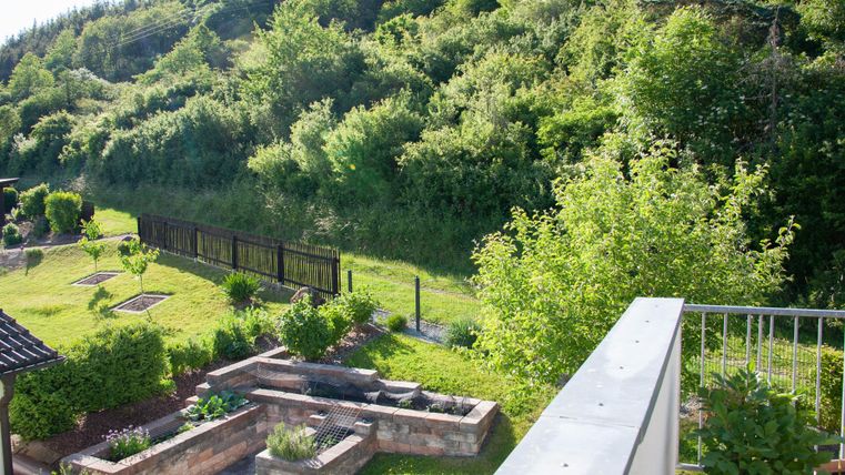 A well-maintained garden with green plants and a small herb bed. In the background, wooded hills and a fence can be seen.