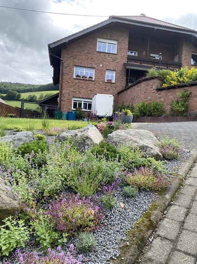 Een huis met bruine bakstenen en een tuin met kleurrijke planten. Op de voorgrond zijn stenen en een geplaveide weg te zien.