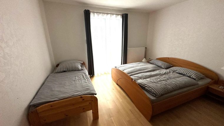 Een eenvoudige slaapkamer met een groot tweepersoonsbed en een eenpersoonsbed. Er is een raam met gordijnen dat daglicht binnenlaat.